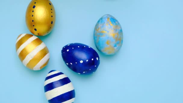复活节彩蛋在滚动,互相敲打着蓝色的桌子.鸡蛋色彩艳丽,经典的蓝色、白色和金色.复活节快乐简约的风格顶部视图 — 图库视频影像