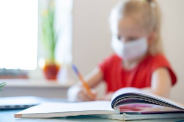 Blurred blonde schoolgirl in medical mask studying at home, doing school homework, writing in notebook. Reading training books on table. coronavirus quarantine. Distance learning online education. clipart