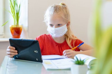 Distance learning online education. Sickness schoolgirl in medical mask studying at home with digital tablet in hand and doing school homework. Training books and notebooks on table. clipart