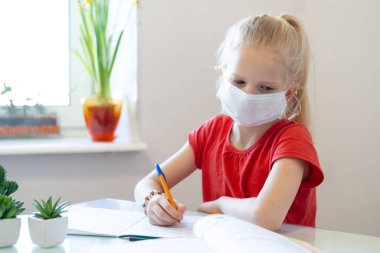 Serios sad blonde schoolgirl in medical mask stydying at home, doing school homework, writing in notebook. Reading training books on table. Back to school. Distance learning online education. clipart