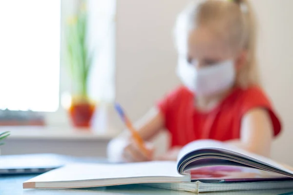 Blurred blonde schoolgirl in medical mask studying at home, doing school homework, writing in notebook. Reading training books on table. coronavirus quarantine. Distance learning online education.