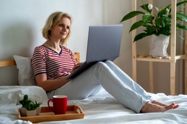 Çevrimiçi eğitim ve iş öğrenme mesafesi. Ofiste çalışan mutlu bir kadın, uzaktan evde, yatakta çalışıyor. Bilgisayar kullanarak ve kahve içerek.