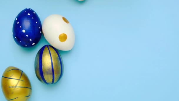 Los huevos de Pascua están rodando, golpeándose unos a otros en la mesa azul. Huevos de moda de color clásico azul, blanco y dorado. Feliz Pascua. Estilo mínimo. Vista superior — Vídeos de Stock