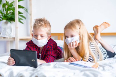 Uzaktan öğrenme çevrimiçi eğitim. Hasta okul çocuğu ve maskeli kız evde dijital tablet dizüstü bilgisayarla ders çalışıyor ve ödev yapıyor. Eğitim kitaplarıyla yatakta oturmak.