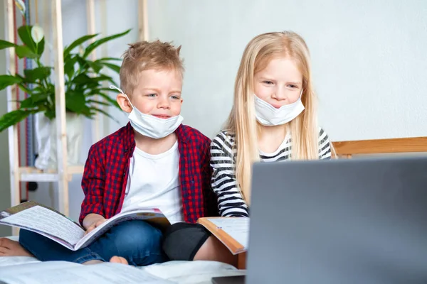 Kształcenie online na odległość. Choroba chłopiec i dziewczyna w masce medycznej studiuje w domu z notebooka laptopa cyfrowego i odrabianie zadań domowych. Siedzenie na łóżku z książkami treningowymi — Zdjęcie stockowe