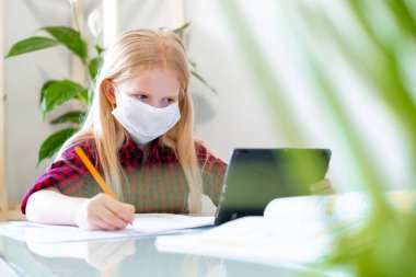 Distance learning online education. Sickness schoolgirl in medical mask studying at home with digital tablet in hand and doing school homework. Training books and notebooks on table. clipart