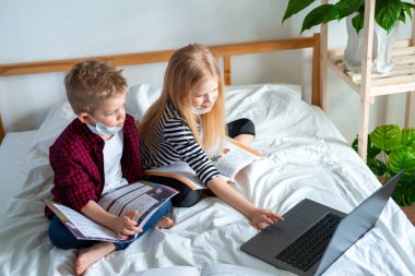 Distance learning online education. Sickness school boy and girl in medical mask studying at home with digital tablet laptop notebook and doing homework. Sitting on bed with training books clipart
