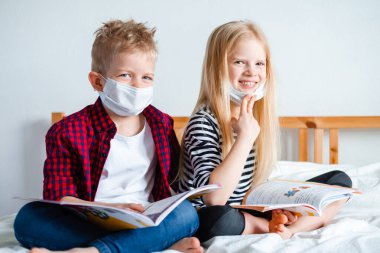 Distance learning online education. Sickness school boy and girl in medical mask studying at home with digital tablet laptop notebook and doing homework. Sitting on bed with training books clipart