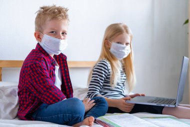 Uzaktan öğrenme çevrimiçi eğitim. Hasta okul çocuğu ve maskeli kız evde dijital tablet dizüstü bilgisayarla ders çalışıyor ve ödev yapıyor. Eğitim kitaplarıyla yatakta oturmak.