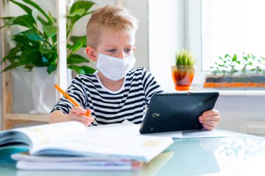 Distance learning online education. Sickness schoolboy in medical mask studying at home with digital tablet in hand and doing school homework. Training books and notebooks on table. clipart