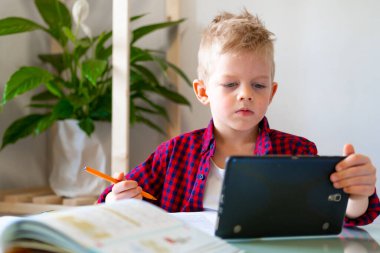 Uzaktan öğrenme çevrimiçi eğitim. Öğrenci evde dijital tabletle ders çalışıyor ve okul ödevi yapıyor. Çalışma kitapları ve defterler masada..