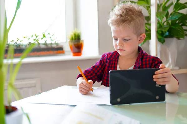 Kształcenie online na odległość. Uczeń uczy się w domu z tabletem cyfrowym i odrabia lekcje. Książki szkoleniowe i zeszyty na stole. — Zdjęcie stockowe