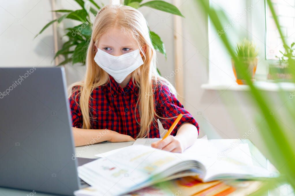 Distance learning online education. Sickness schoolgirl in medical mask studying at home with digital tablet in hand and doing school homework. Training books and notebooks on table.