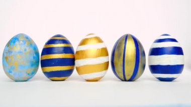 Paskalya yumurtaları yuvarlanıyor, birbirlerini beyaz masaya vuruyorlar. Yumurtaların modası geçmiş klasik mavi, beyaz ve altın rengi. Mutlu Paskalyalar. Asgari biçim. 