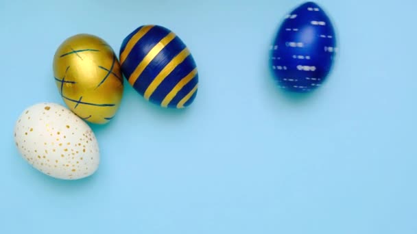 Los huevos de Pascua están rodando, golpeándose unos a otros en la mesa azul. Huevos de moda de color clásico azul, blanco y dorado. Feliz Pascua. Estilo mínimo. Vista superior — Vídeo de stock