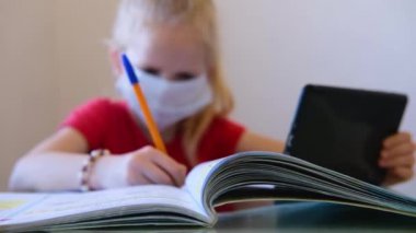 Tıp maskeli, bulanık sarışın kız evde ders çalışıyor, okul ödevi yapıyor, deftere yazıyor. Masada eğitim kitapları okuyor. Okula dönelim. Uzaklık öğrenimi çevrimiçi eğitim.