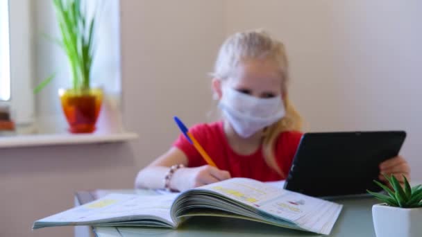 Educación a distancia en línea. Colegiala enferma en máscara médica estudiando en casa con tableta digital en la mano y haciendo la tarea escolar. Libros de entrenamiento y cuadernos en la mesa . — Vídeos de Stock
