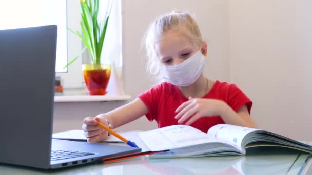 Educación a distancia en línea. Colegiala enferma en máscara médica estudiando en casa con tableta digital en la mano y haciendo la tarea escolar. Libros de entrenamiento y cuadernos en la mesa . — Vídeos de Stock