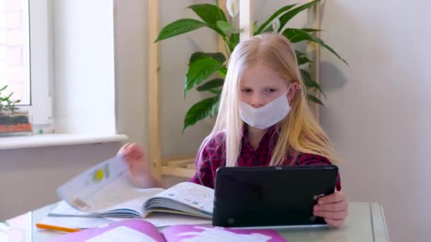 Educación a distancia en línea. Colegiala enferma en máscara médica estudiando en casa con tableta digital en la mano y haciendo la tarea escolar. Libros de entrenamiento y cuadernos en la mesa . — Vídeos de Stock