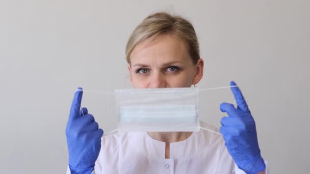 La doctora rubia cirujana en uniforme blanco y guantes azules se pone máscara médica. Concepto de higiene, tratamiento, medicina — Vídeos de Stock