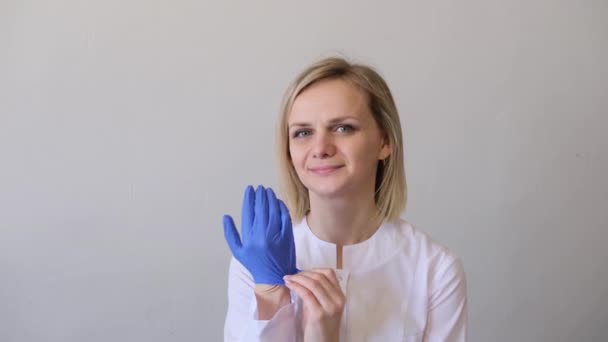 Médica cirurgiã loira de uniforme branco coloca luvas azuis e máscara médica. Conceito de higiene, tratamento, medicina — Vídeo de Stock