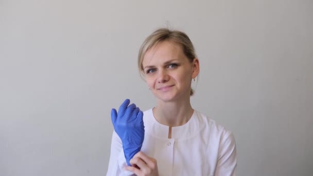 Blondynka lekarz chirurg w białym mundurze zakłada niebieskie rękawiczki i maskę medyczną. Koncepcja higieny, leczenia, medycyny — Wideo stockowe
