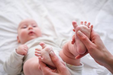 Bebek ayakları annenin ellerinde. Kafkasyalı genç bir kadın, beyaz yatakta mutlu bir bebeğe masaj yapıyor. Bebek bakımı, spor ve mutlu annelik.