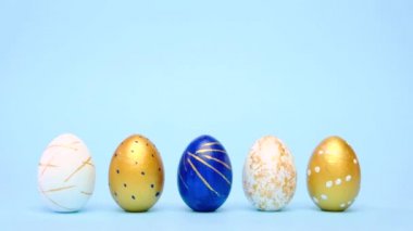 Paskalya yumurtaları yuvarlanıyor, birbirlerini mavi masaya vuruyorlar. Yumurtaların modası geçmiş klasik mavi, beyaz ve altın rengi. Mutlu Paskalyalar. Asgari biçim. 