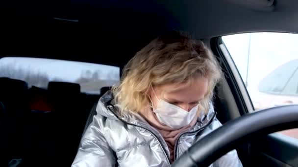 Conductor rubio rizado de la muchacha de 4k que pone en dos máscara médica y guantes protectores azules en coche antes de conducir. Mujer con chaqueta caliente contra el resfriado gripe común. Quédate en casa en cuarentena . — Vídeo de stock