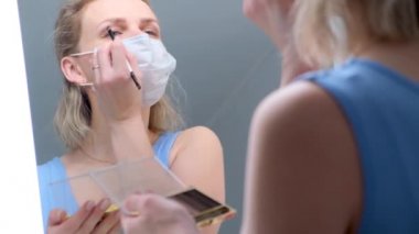 4 bin. Tıbbi koruyucu maskeli sarışın kız göz makyajı yapıyor, göz farı sürüyor. Mirrow 'a benziyor. Soğuk algınlığına karşı kadın. Evde kalıp karantina altında kalın.