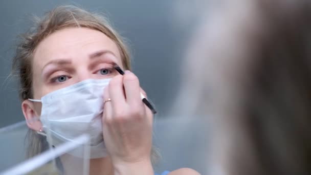 4k. Blond flicka i medicinsk skyddsmask gör ögonmakeup, applicerar ögonskugga. Titta i spegeln. Kvinna mot förkylning vanlig influensa. Stanna hemma karantän. — Stockvideo