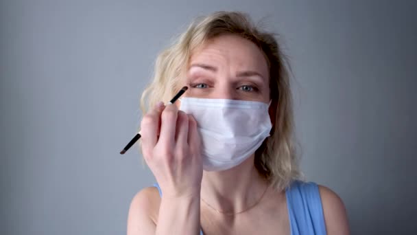 4k医療保護マスクのCurlyブロンド喜びの女の子は、アイメイクを行い、アイシャドウを適用します。カメラを見て。風邪の一般的なインフルエンザに対する女性。隔離室にいて. — ストック動画