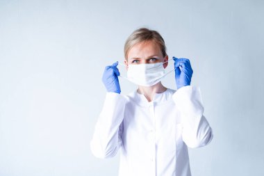 Beyaz üniformalı sarışın kadın doktor, gri üzerine tıbbi maske takan koruyucu mavi eldivenler. Sağlık hizmeti, evde kal. Kopyalama alanı