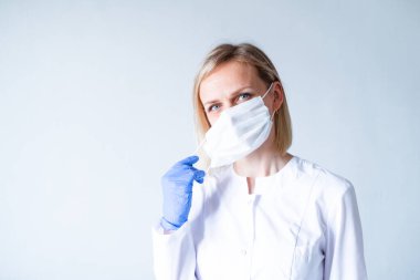 Beyaz üniformalı sarışın kadın doktor, gri üzerine tıbbi maske takan koruyucu mavi eldivenler. Sağlık hizmeti, evde kal. Kopyalama alanı