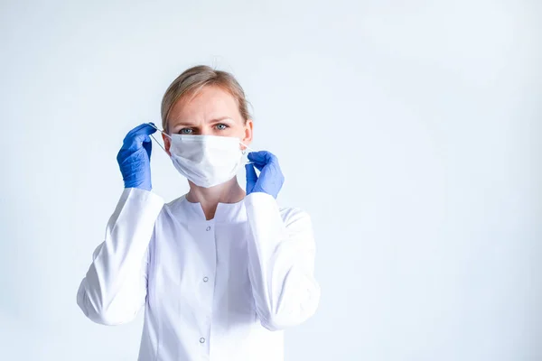 Blonde female doctor surgeon in white uniform, protective blue gloves putting on medical mask over grey. Healthcare, stay home. copyspace — Stock Photo, Image