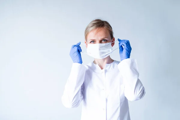 Blonde female doctor surgeon in white uniform, protective blue gloves putting on medical mask over grey. Healthcare, stay home. copyspace — Stock Photo, Image