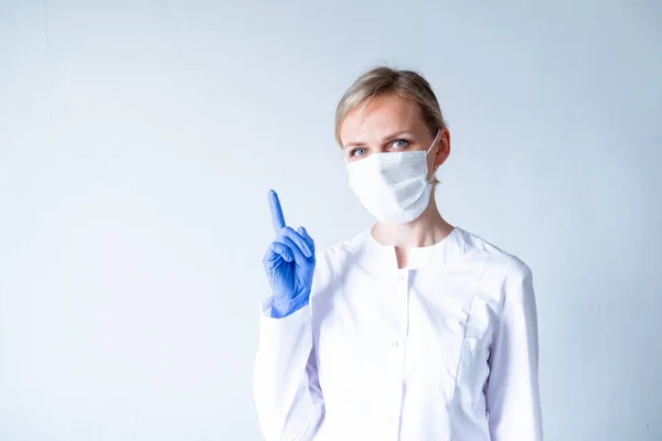Attention. Blonde female doctor surgeon in white uniform, protective blue gloves and medical mask showing finger up over grey. — Stock Photo, Image