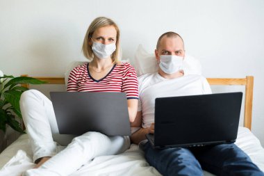 Genç aile evden uzakta bilgisayarın başında yatağında çalışıyor. Tıbbi maskeli karantina altındaki bir çift koronavirüs. Kendine dikkat et. Mesafe öğrenme, eğitim ve çalışma. İnternetten gıda siparişi