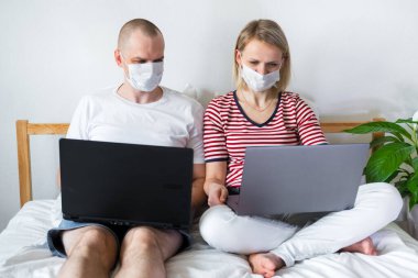 Genç aile evden uzakta bilgisayarın başında yatağında çalışıyor. Tıbbi maskeli karantina altındaki bir çift koronavirüs. Kendine dikkat et. Mesafe öğrenme, eğitim ve çalışma. İnternetten gıda siparişi
