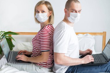 Aile uzaktan çalışıyor, arka arkaya yatakta oturuyorlar. Karantinaya alınmış tıbbi maskeli bir çift koronavirüs. Evde güvende kalın. Mesafe öğrenme, eğitim ve çalışma. İnternetten gıda siparişi