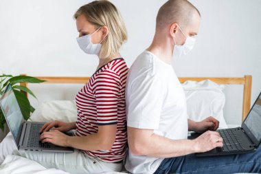 Aile uzaktan çalışıyor, arka arkaya yatakta oturuyorlar. Karantinaya alınmış tıbbi maskeli bir çift koronavirüs. Evde güvende kalın. Mesafe öğrenme, eğitim ve çalışma. İnternetten gıda siparişi