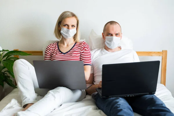 young family works remotely from home on bed at the computer. Quarantined couple coronavirus in medical masks. Stay home safe. Distance learning, education and work. Order food products online