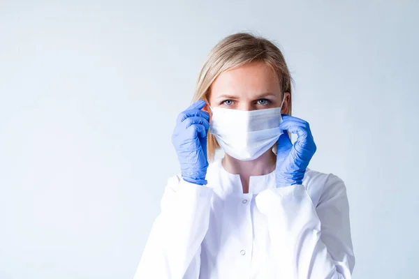 Blonde female doctor surgeon in white uniform, protective blue gloves putting on medical mask over grey. Healthcare, stay home. copyspace — Stock Photo, Image