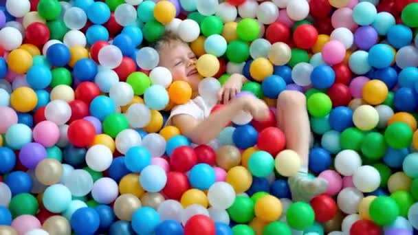 Blonďatý chlapec rozhazuje míčky po různých stranách, leží na vícebarevných plastových kuličkách ve velkém suchém brouzdališti v hracím centru. Usmívá se na kameru. Bavte se v herně. Volnočasová činnost — Stock video