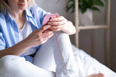 Akıllı telefon kullanan sarışın bir kadın yatakta oturuyor. Haberleri ve online kitapları okumak. Sosyal medya, instagram, evde kalıp karantina