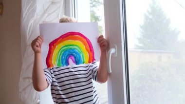 4K. Evde Covid-19 karantinası sırasında küçük sarışın çocuk gökkuşağı deseninin arkasına saklanarak eğleniyor. Umarım her şey yolunda gider. Gökkuşağını kovala