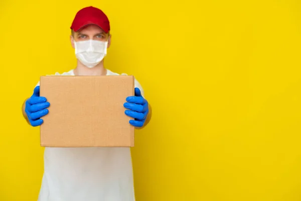 Delivery man in red cap white t-shirt uniform face medical mask gloves hold empty cardboard box on yellow studio background. Service coronavirus. Online shopping. mock up. copyspace
