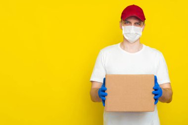 Kırmızı şapkalı beyaz tişörtlü teslimatçı. Yüzü tıbbi maske eldivenli. Sarı stüdyo arka planında boş karton kutu var. Hizmet koronavirüsü. İnternetten alışveriş. Model yap. kopyalama alanı