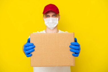 Kırmızı şapkalı beyaz tişörtlü teslimatçı. Yüzü tıbbi maske eldivenli. Sarı stüdyo arka planında boş karton kutu var. Hizmet koronavirüsü. İnternetten alışveriş. maket yap