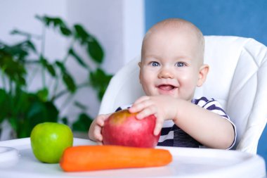 Bebek maması. Büyük kırmızı elmalı ve sandalyesinde havuç olan gülümseyen beyaz bir çocuk. Sağlıklı yemek. Taze sebzeler ve meyveler. Yeni doğmuş bebekler.